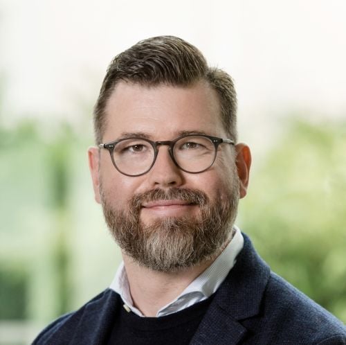 Portrait of a man with glasses and a beard wearing a dark blazer against a softly blurred background