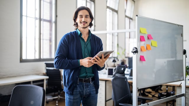 Un formateur USU IT Monitoring se tient dans un bureau moderne, tenant une tablette et souriant avec assurance. Derrière lui, un tableau blanc avec des notes autocollantes colorées indique une session de formation ou de planification active dans le domaine de l'IT Monitoring.