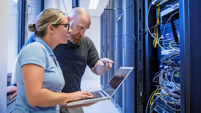 Deux informaticiens travaillent ensemble dans une salle de serveurs dans le domaine de la surveillance informatique. Une femme tient un ordinateur portable tandis qu'un homme pointe l'écran. En arrière-plan, plusieurs racks de serveurs avec des câbles de réseau et du matériel informatique sont visibles.