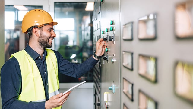 Un technicien équipé d'un casque de protection et d'une tablette surveille des installations de commutation dans la salle de contrôle. Symbolise la surveillance de l'infrastructure des centres de données et des bâtiments.