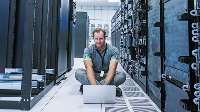 Un informaticien est assis avec son ordinateur portable dans la salle des serveurs devant de grands racks de serveurs. Symbolise la surveillance des ordinateurs centraux.