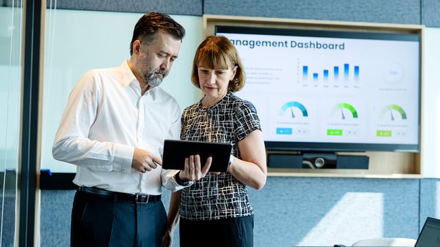 Deux cadres analysent les données de capacité sur une tablette, tandis qu'un tableau de bord de gestion s'affiche en arrière-plan. Symbolise la gestion de la capacité