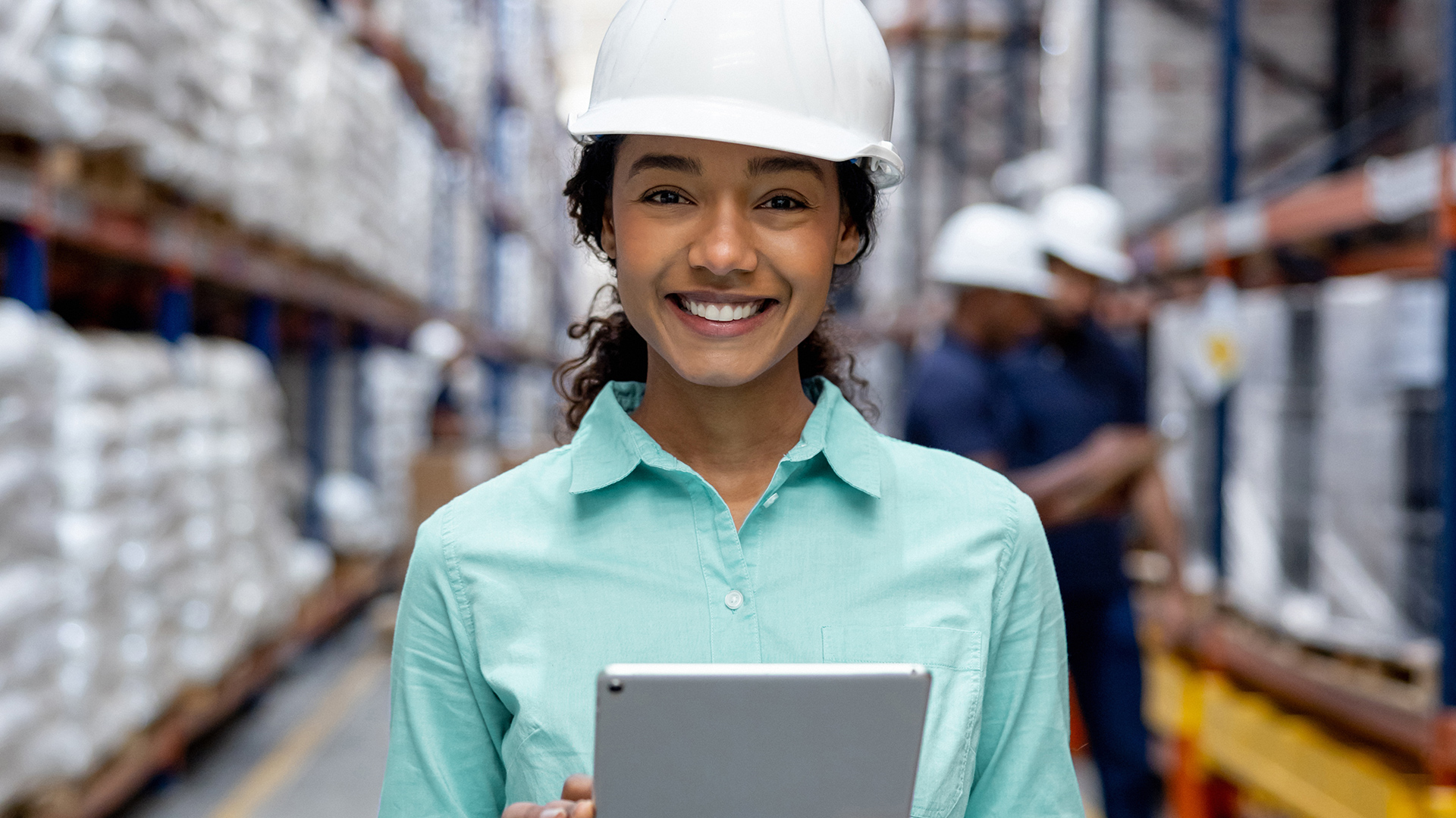 A woman wearing a helmet smiling in a production hall with a tablet in her hand.